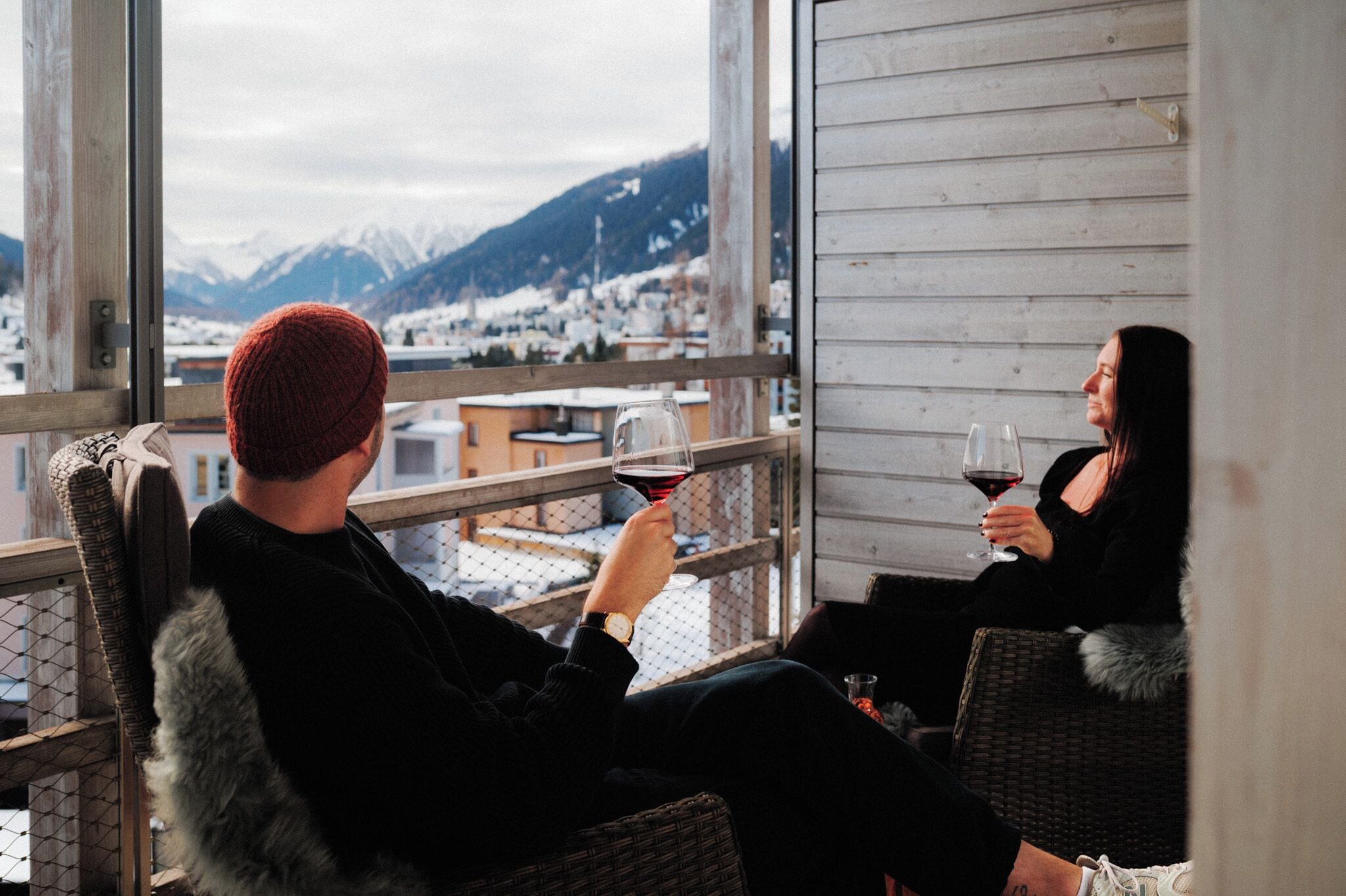 Two people seated on wicker chairs on a balcony with a view of a snowy mountain town.