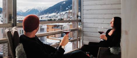 Two people seated on wicker chairs on a balcony with a view of a snowy mountain town.