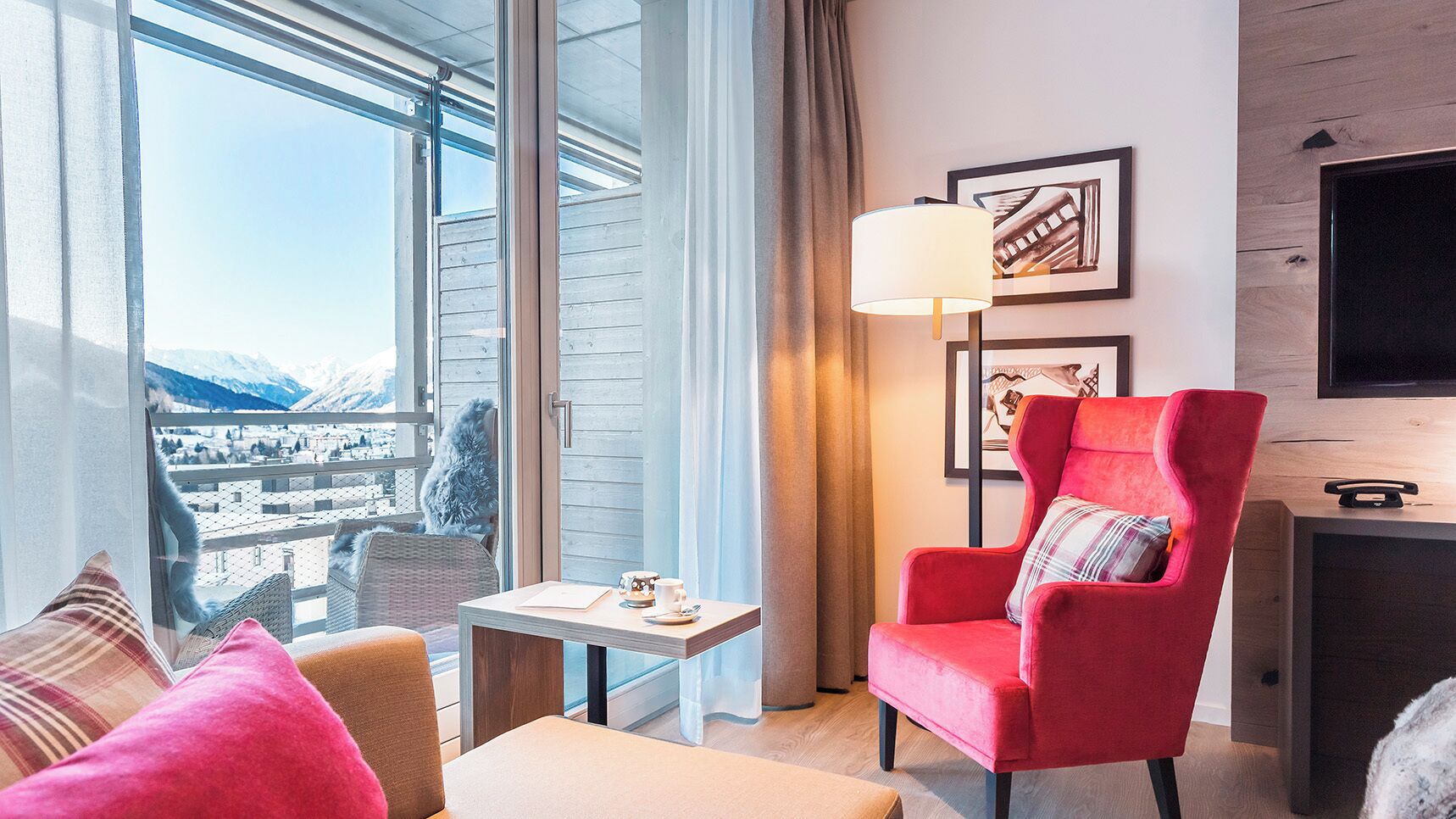 Una habitación de hotel moderna con un ventanal grande, un sillón rojo, una mesita y un televisor.