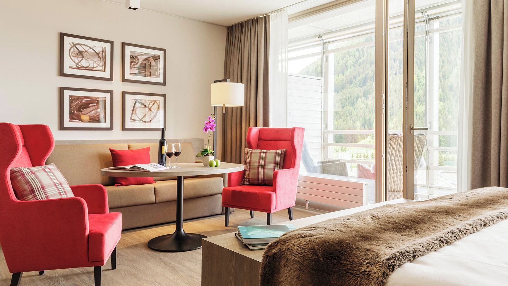Habitación de hotel con una cama, dos sillones rojos, un sofá, una mesa con lámpara y ventana con vistas a árboles.