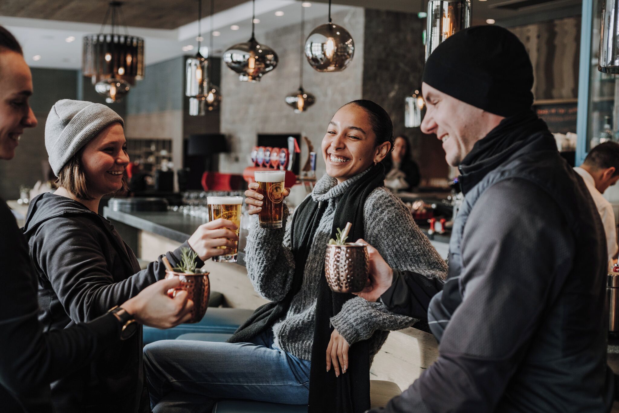 Un groupe de personnes est assis et trinque avec des boissons dans un cadre intérieur moderne, éclairé par des suspensions.