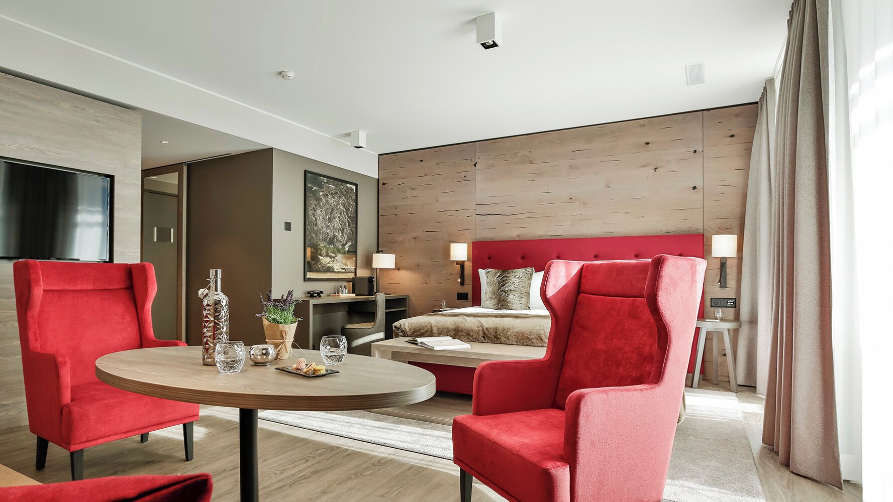Quarto de hotel moderno com uma cama, duas poltronas vermelhas, uma mesa redonda com um vaso e copos, e uma janela grande com cortinas.