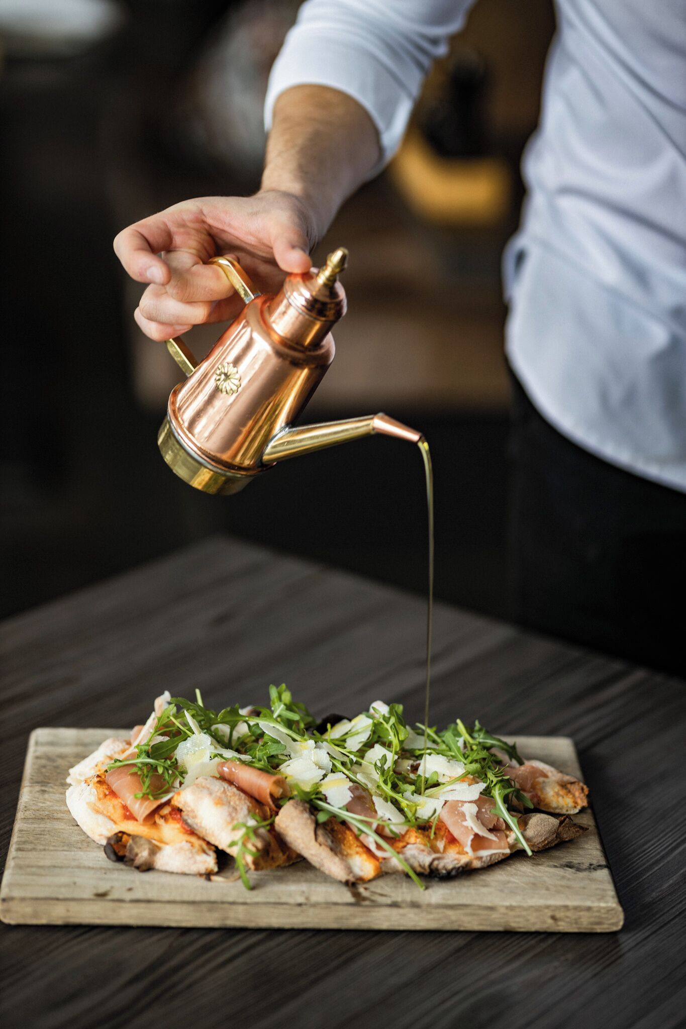 Une personne verse un liquide d’un pichet en cuivre sur une pizza posée sur une planche en bois.