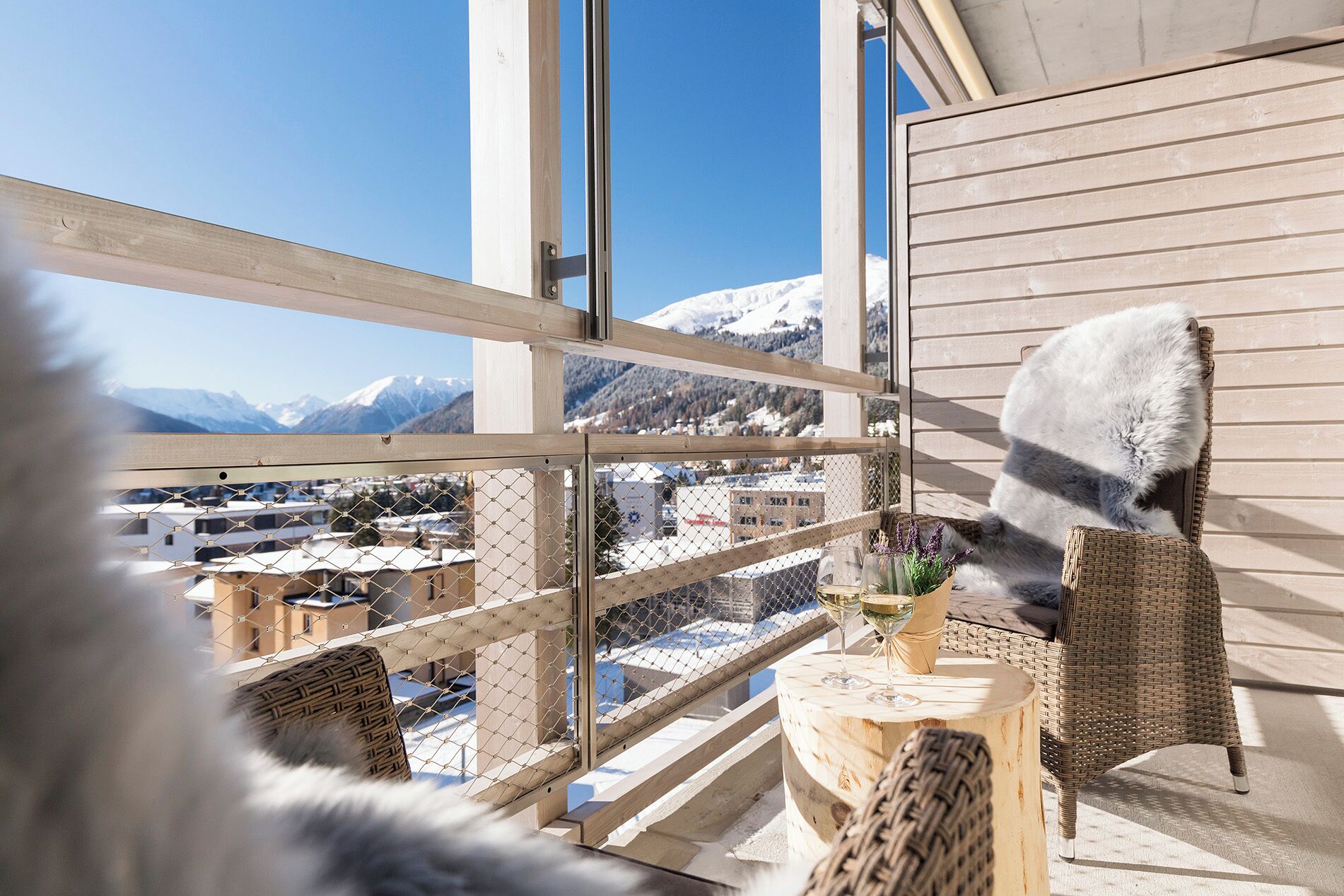 A high-rise room with a view of snow-capped mountains, wicker furniture, and a glass table with a drink.