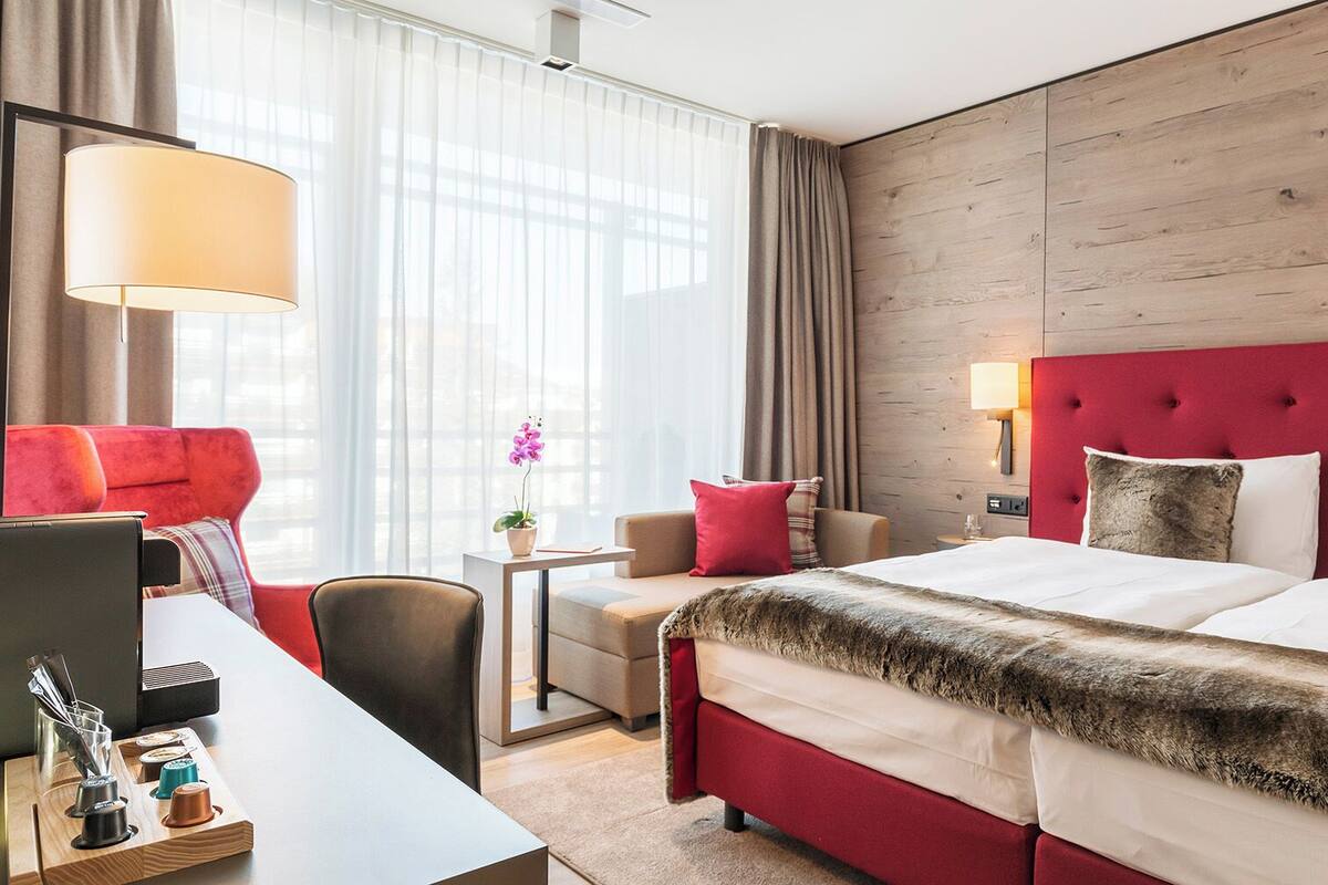 Ein Hotelzimmer mit einem Bett, einem Schreibtisch mit Laptop, einem Sessel, einem kleinen Tisch mit Topfpflanze und einem Fenster mit Vorhängen.