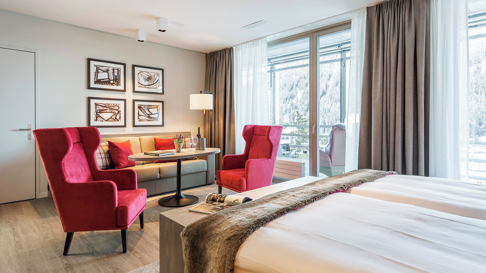 A hotel room with a bed, two red armchairs, a sofa, a small table with a lamp, and framed artwork on the wall.