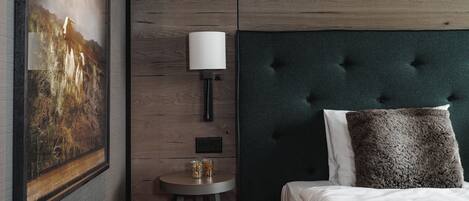A bedroom with a tufted headboard, a framed landscape painting, a bedside lamp, and a small table with glasses.