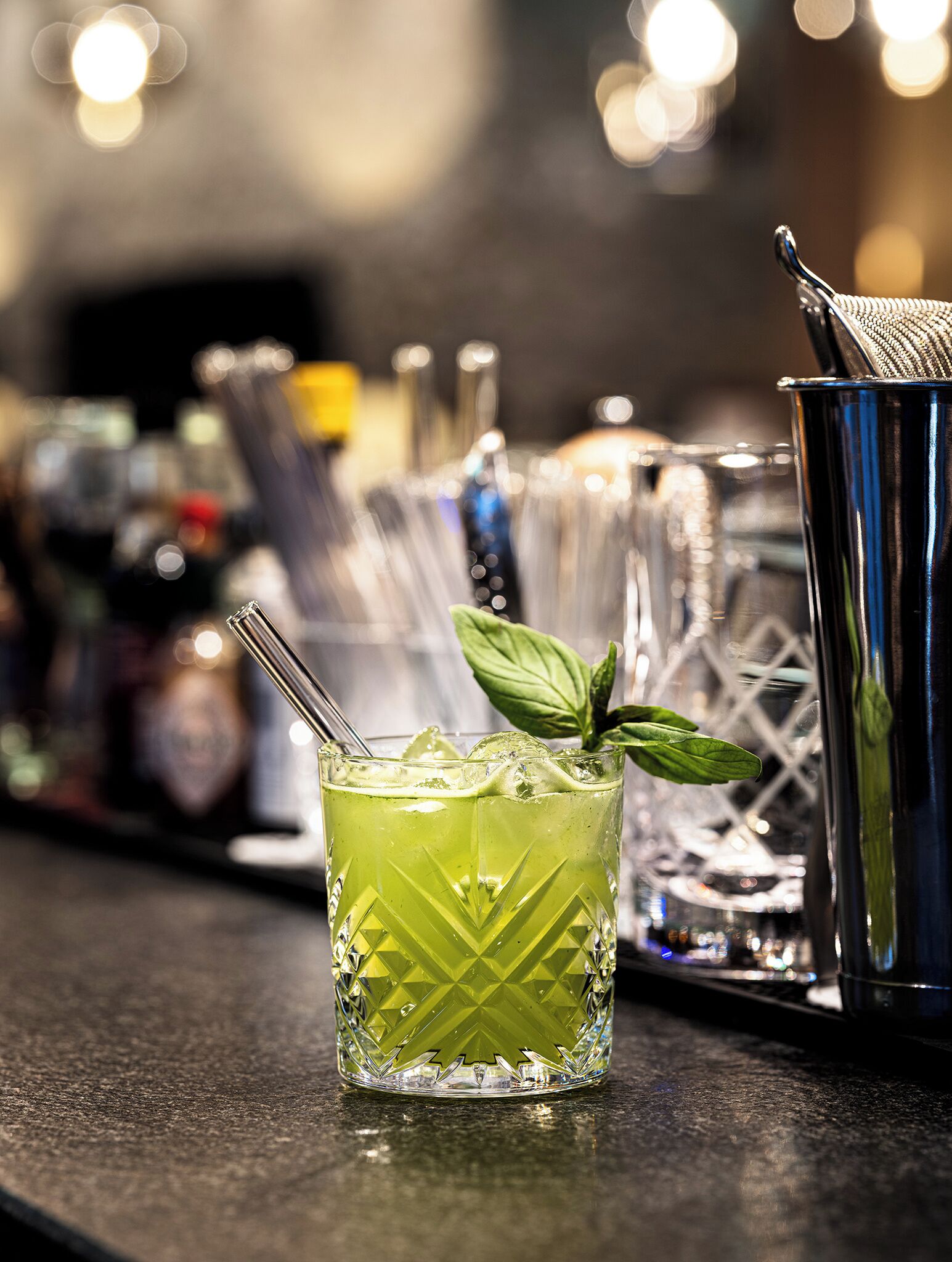 Un bar con un cóctel verde, un coctelera metálica y un vaso adornado con menta.