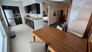 A compact kitchen with a refrigerator, microwave, and dark cabinets. There's a dining area with a wooden table and a single chair.