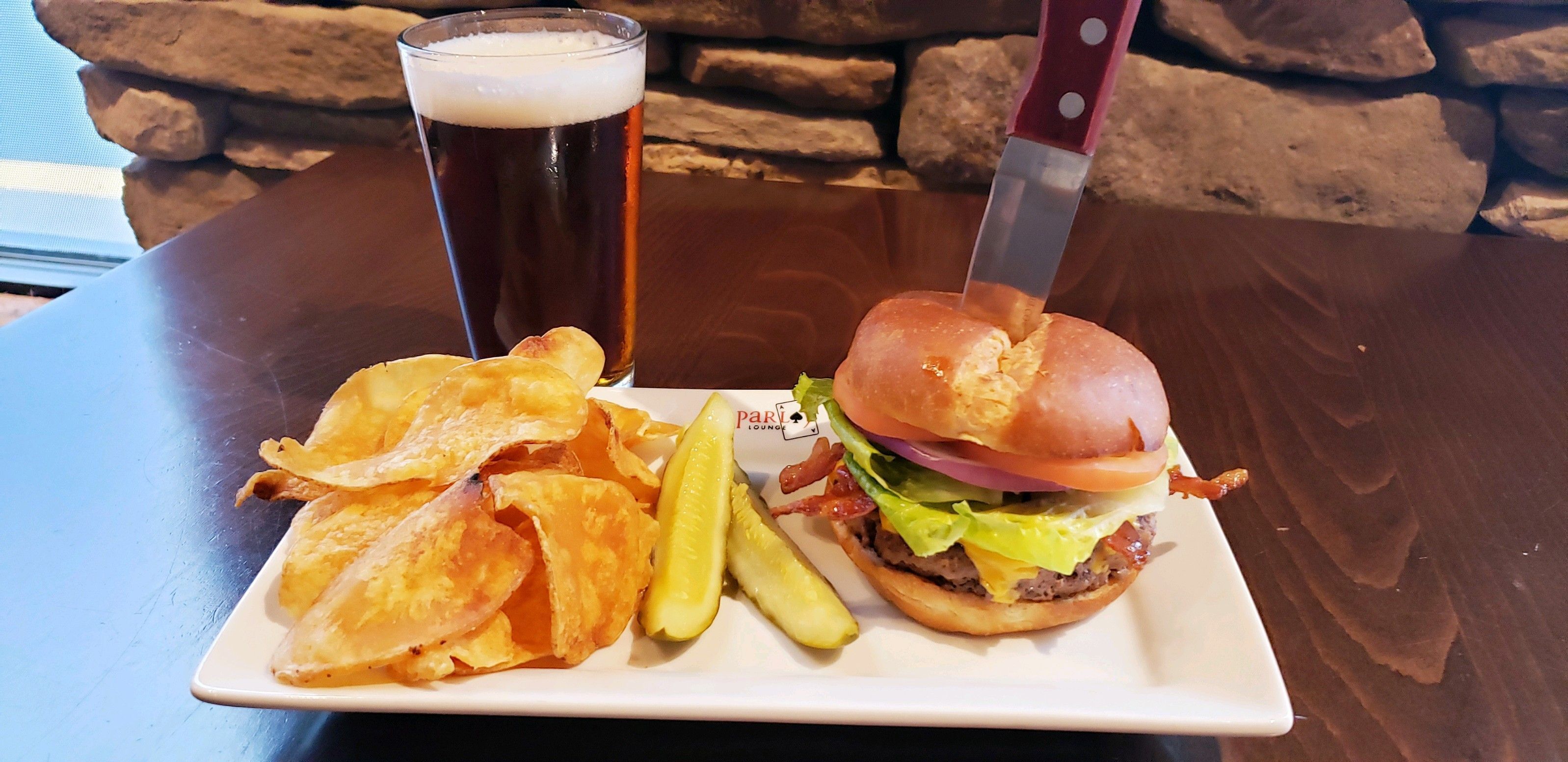 Un repas composé d’un hamburger, de frites et d’un cornichon, servis sur une assiette blanche, avec un verre de bière et un couteau posés sur la table.