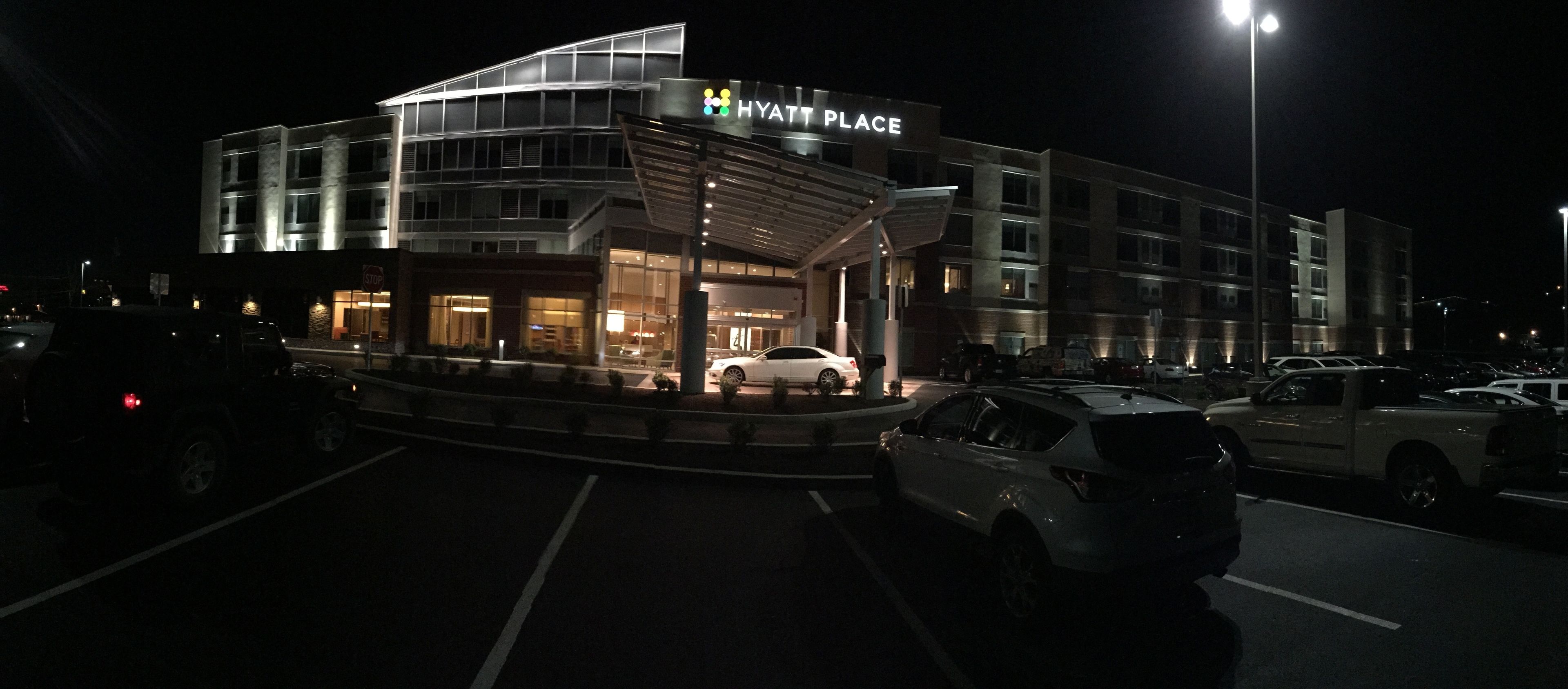A multi-story building with a modern design, labeled "HYATT PLACE," featuring a prominent entrance with a covered area and a parking lot with several cars.