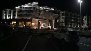 A multi-story building with a modern design, labeled "HYATT PLACE," featuring a prominent entrance with a covered area and a parking lot with several cars.