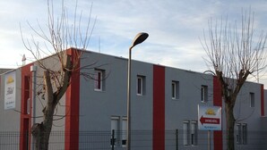 A modern hotel building with a red and grey facade, a fenced parking area, and a street lamp. - Hôtel Les Beaux Soleils (Osny)
