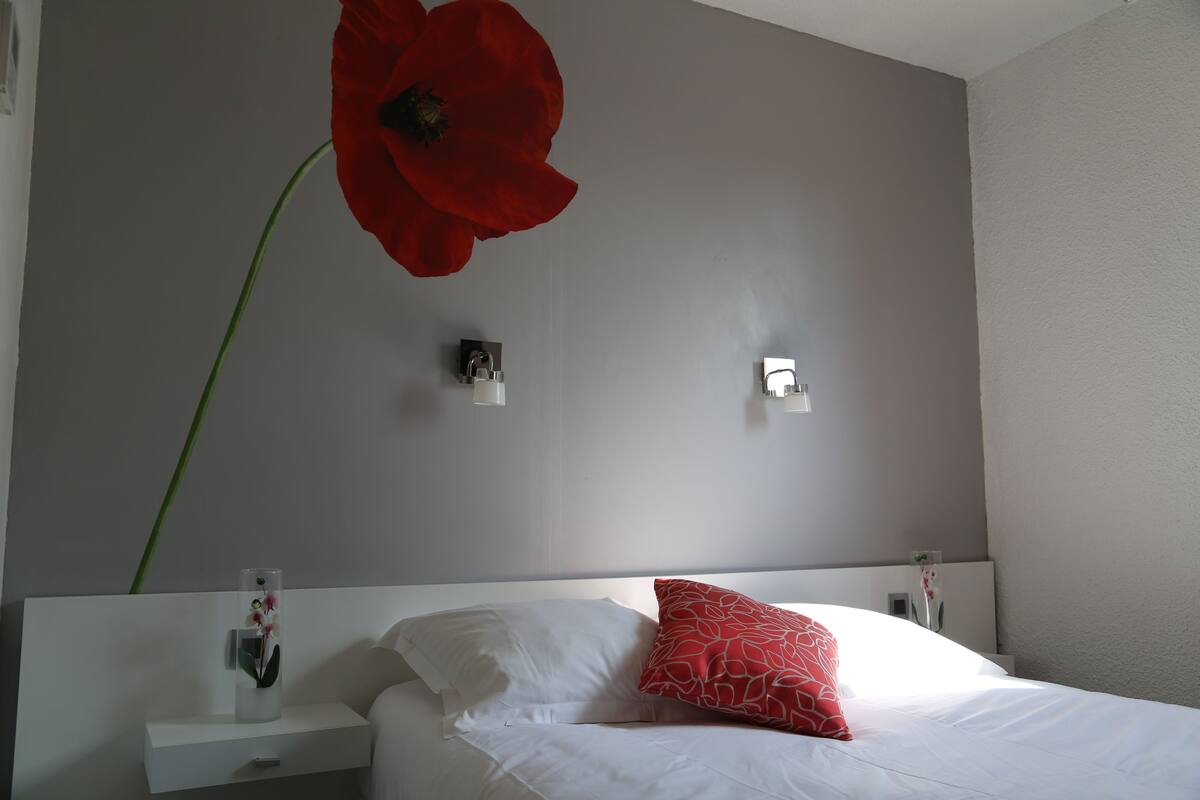 Une chambre à coucher avec un lit, des oreillers blancs, une décoration florale rouge, une table de chevet avec un vase et des lampes fixées au mur.