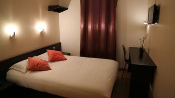 A hotel room with a bed, two red pillows, a nightstand, a desk with a chair, a TV, and a curtain.