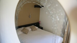 A neatly made bed with white and beige linens, a small bedside table with a lamp, and a wall-mounted light fixture. - Hôtel Les Beaux Soleils (Osny)