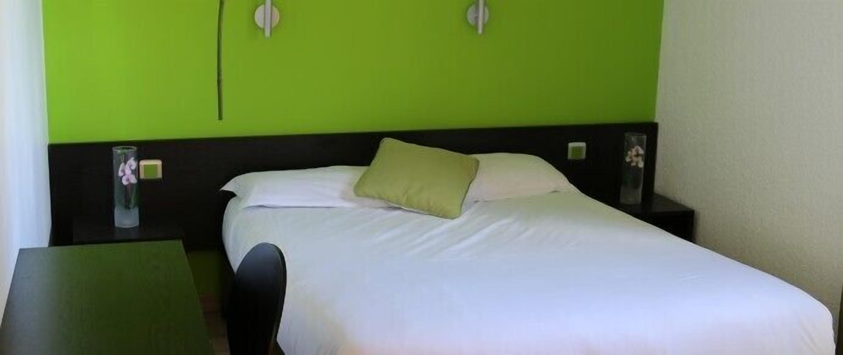 Una cama bien tendida con sábana blanca y una sola almohada verde en una habitación con paredes verdes y un cabecero negro.
