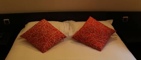 A neatly made bed with white sheets and two red pillows, two wall-mounted light fixtures, and a dark headboard.