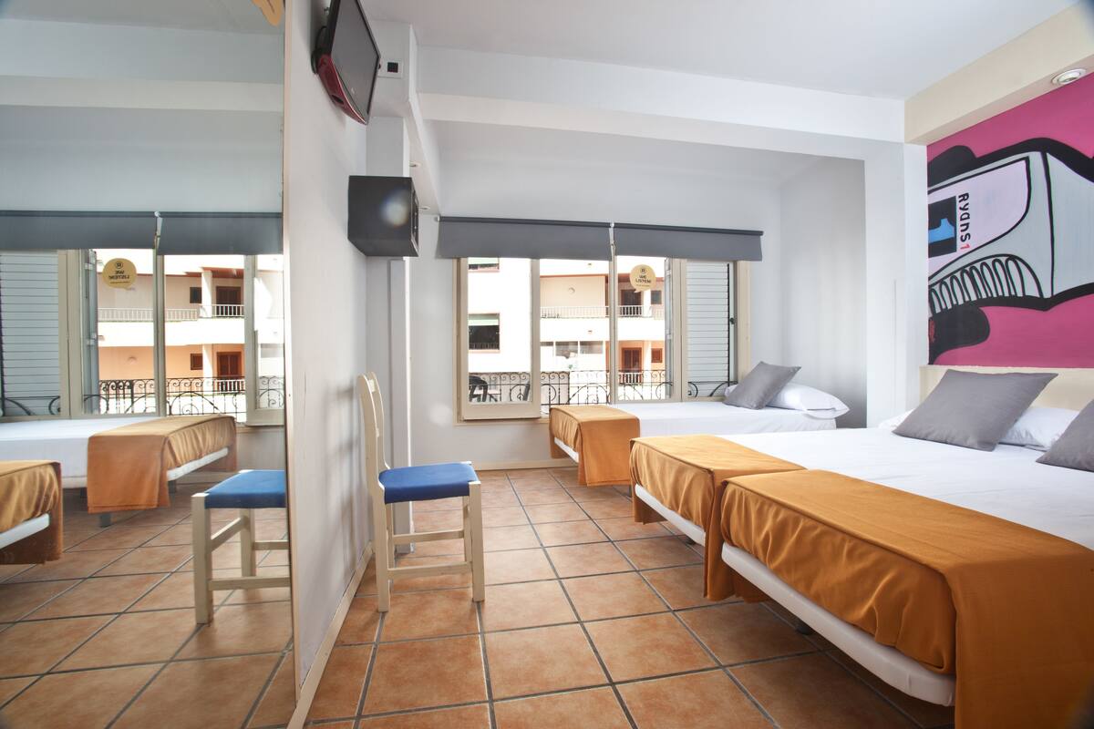 Ein Hotelzimmer mit zwei Betten, einem Sessel, einem kleinen Tisch und einem Fenster mit Blick auf die Außenansicht des Gebäudes.
