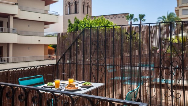 A balcony with a table set for two, a view of a building with a bell tower, and a pool in the background. - Ten Hostel (Ibiza Town)