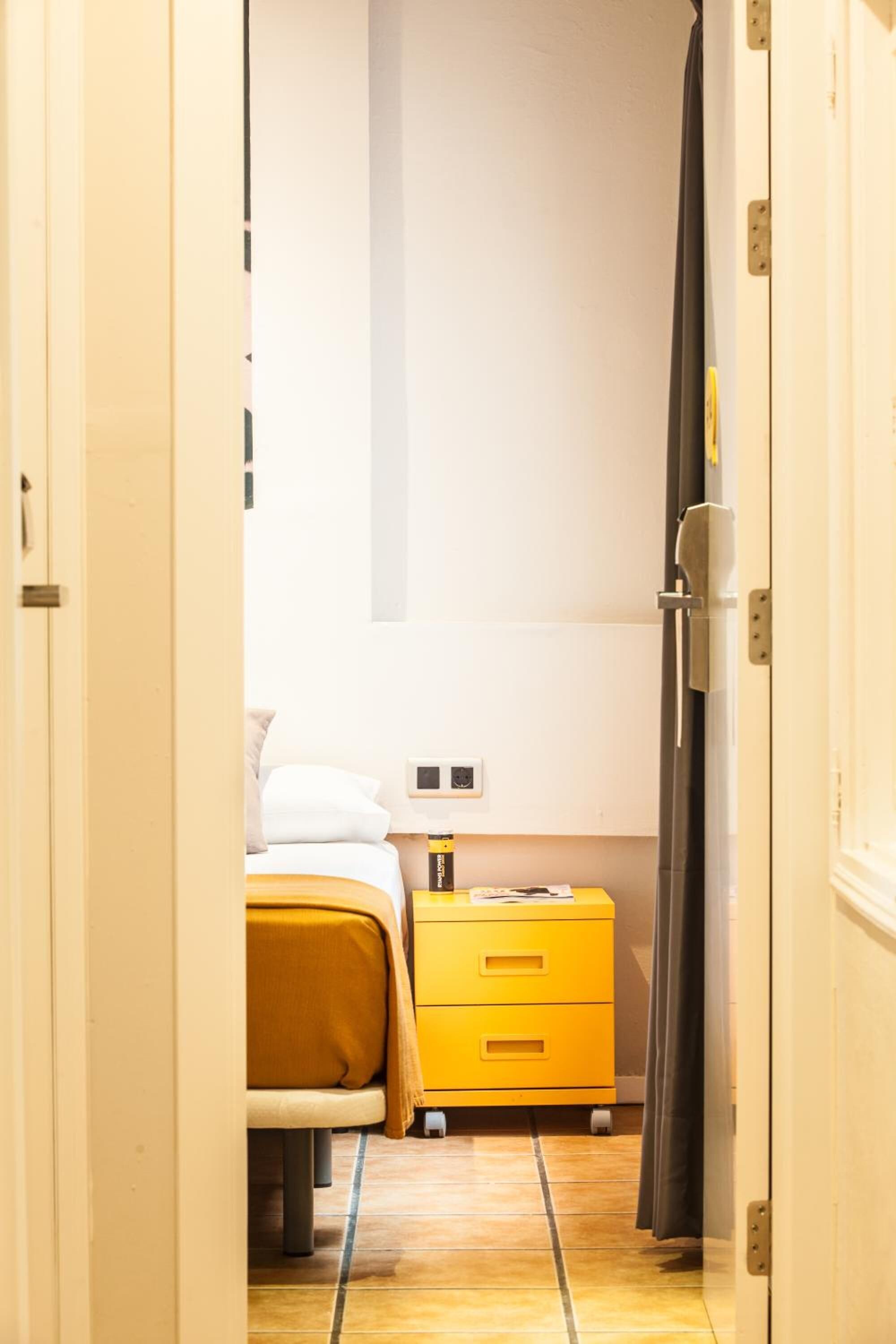 Un pasillo estrecho que conduce a un dormitorio con una cama, una mesita de noche y un aparador amarillo.