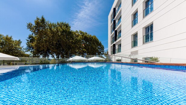 A clear blue pool with a tiled floor, surrounded by a white building with multiple windows and a tree in the background. - Lux Fátima Park - Hotel, Suites & Residence (Ourem)