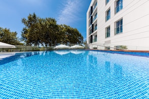 Uma piscina azul límpida com piso de azulejos, cercada por um edifício branco com várias janelas e uma árvore ao fundo.