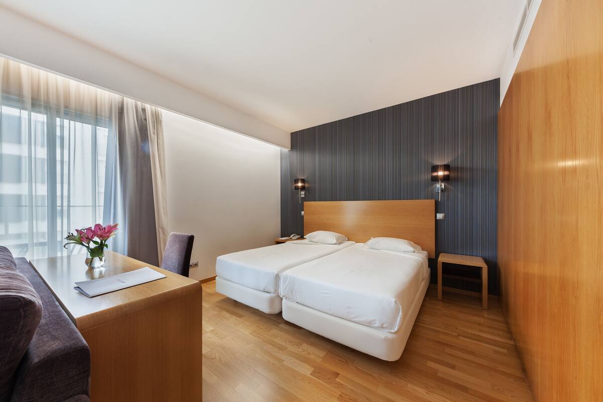 Une chambre d’hôtel avec un lit double, des tables de chevet, un bureau avec une chaise, un petit tabouret et une fenêtre avec des rideaux.