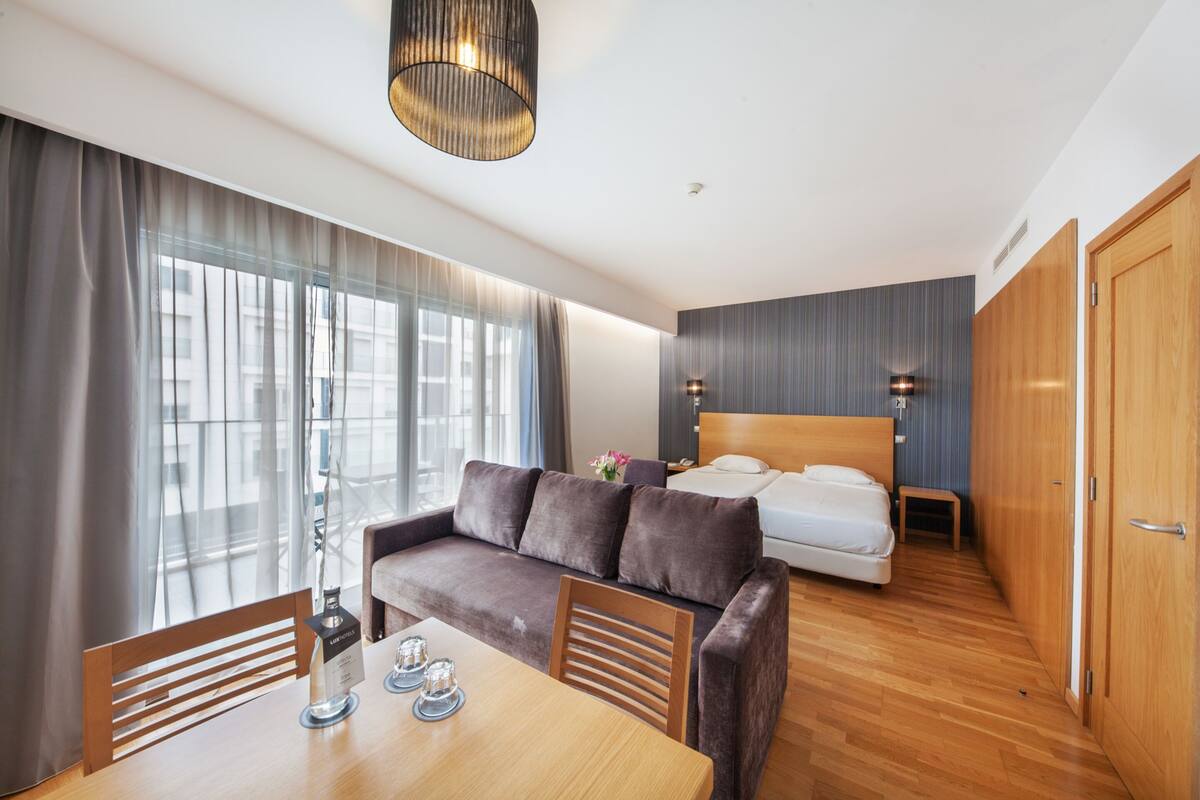 Une chambre d’hôtel moderne avec un plancher en bois, un canapé, un lit, une table à manger et une grande fenêtre avec des rideaux.