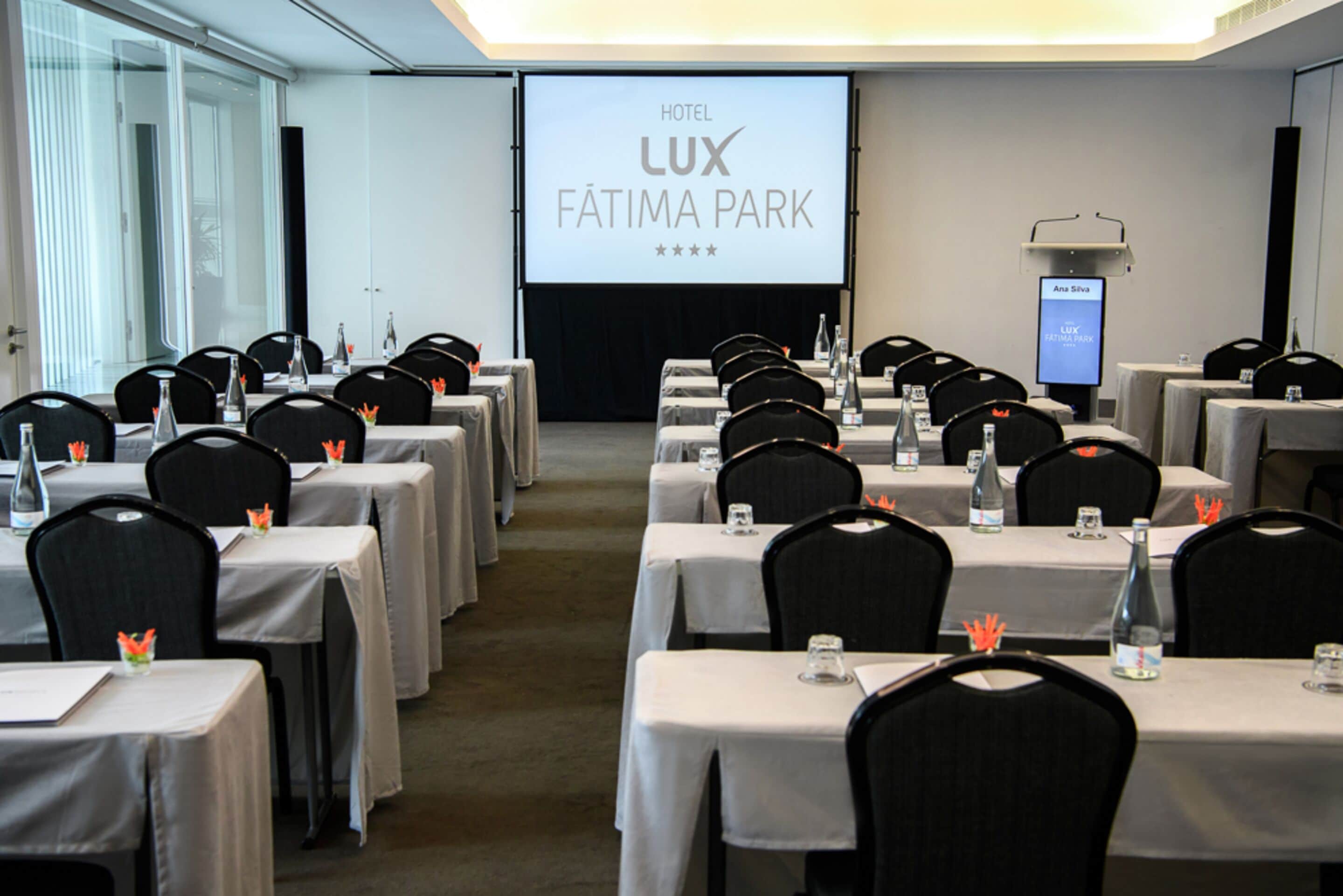 A hotel conference room with tables set for a meeting, a projection screen displaying the hotel name, and a digital display showing a name.