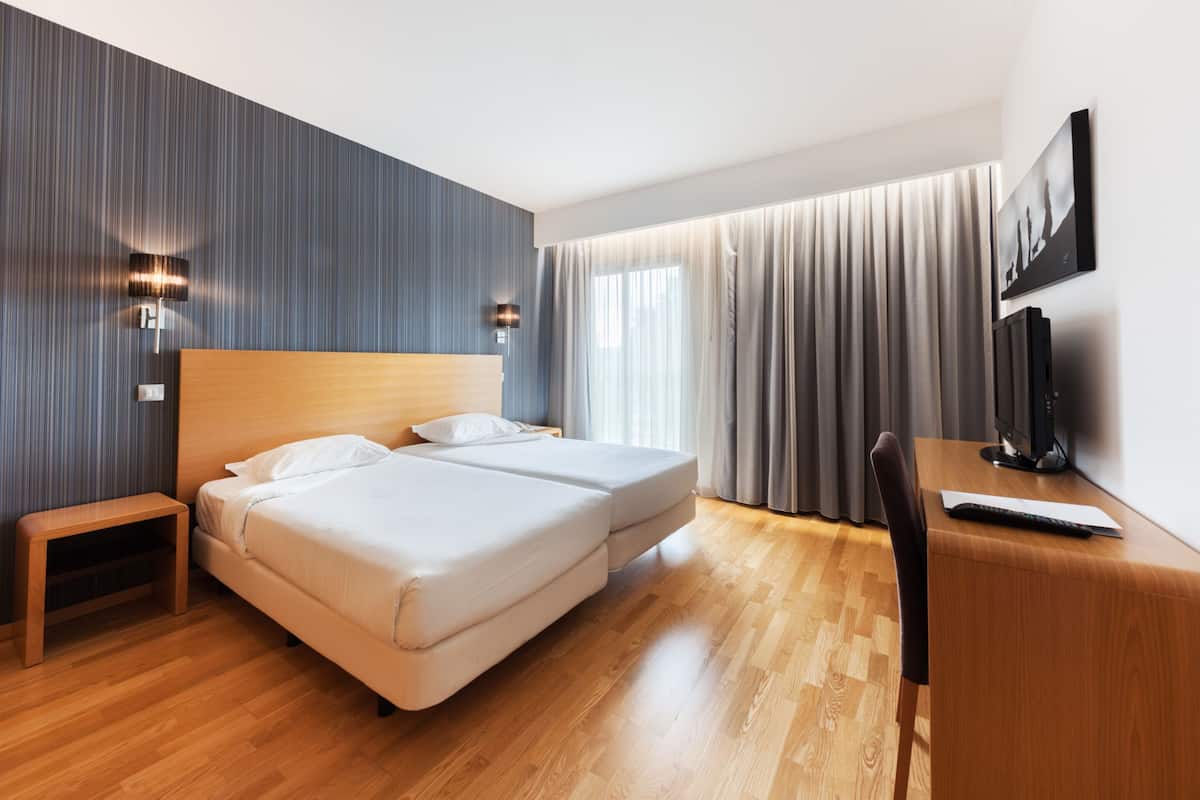 Una habitación de hotel moderna con piso de madera, dos camas individuales, un escritorio con silla, un televisor y una obra de arte colgada en la pared.