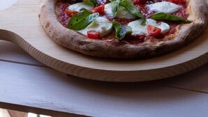Pizza på en træplanke med et glas vand og en potteplante i baggrunden.