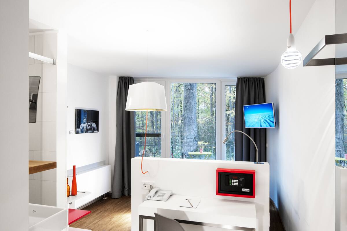 A modern hotel room with a desk, a telephone, a digital control panel, a television, and large windows offering a forest view.