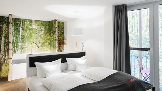 A modern hotel room with a bed, a nightstand, a lamp, a window with curtains, and a view of trees. - V-Hotel (Bonn)