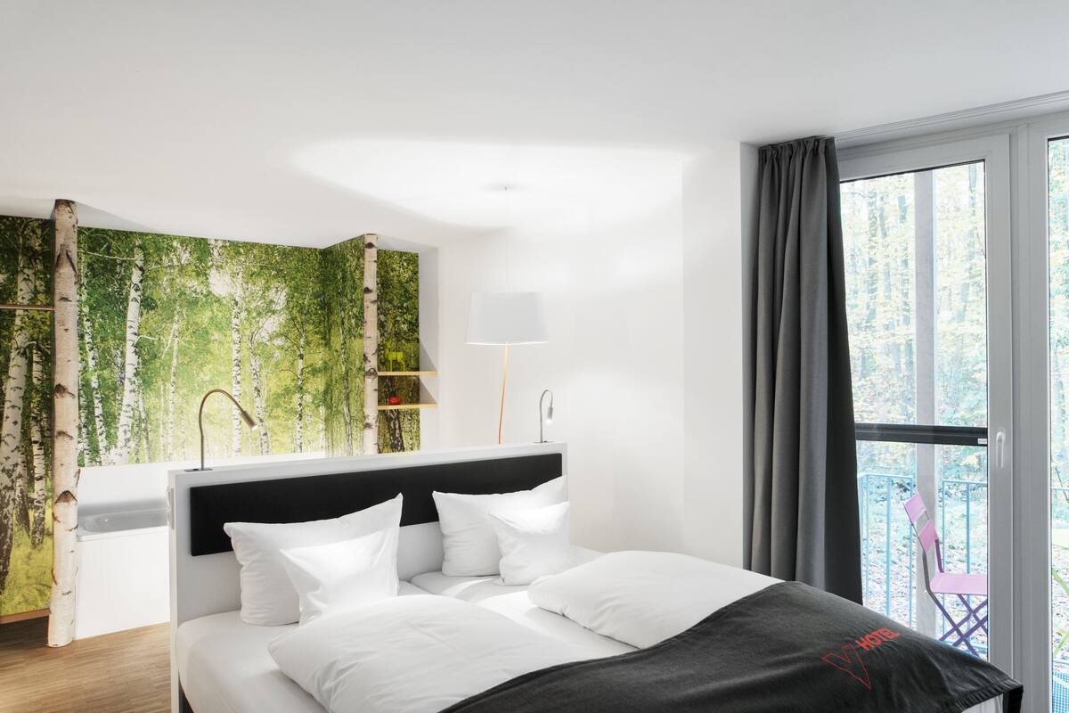 A modern hotel room with a bed, a nightstand, a lamp, a window with curtains, and a view of trees.