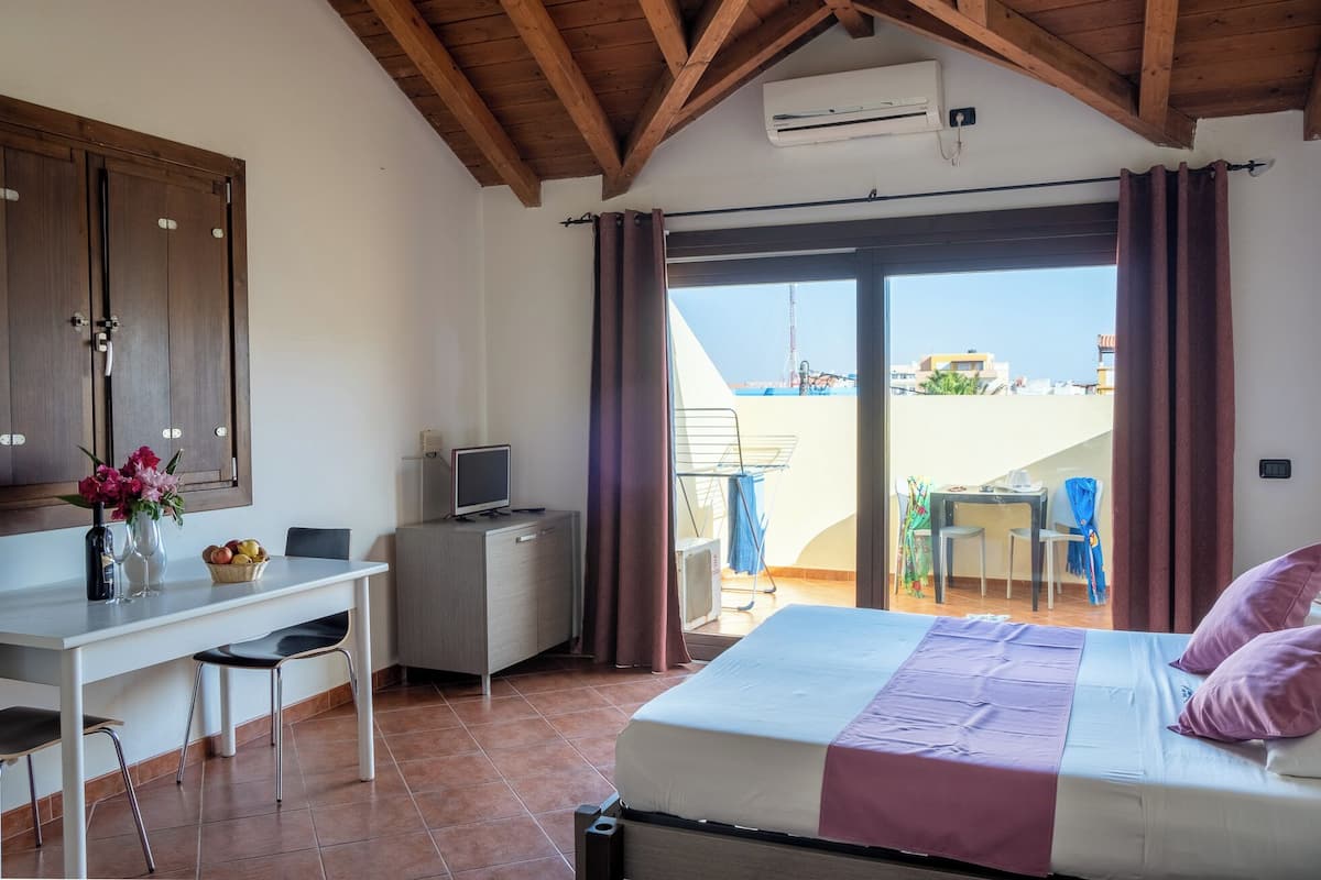 A bedroom with a bed, a desk with a computer, a TV, and a view of the outside.