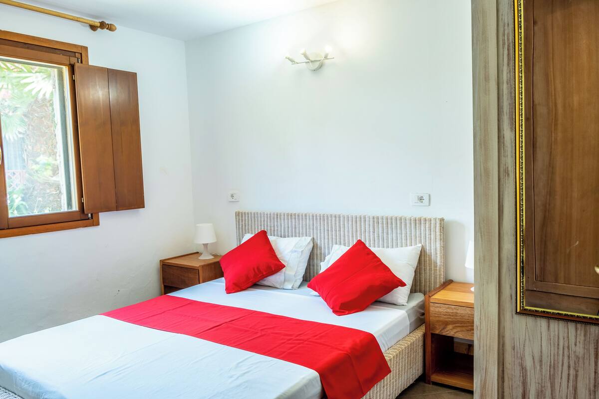 A bedroom with a bed, two bedside tables, a nightstand, a window, and a wall-mounted light fixture.