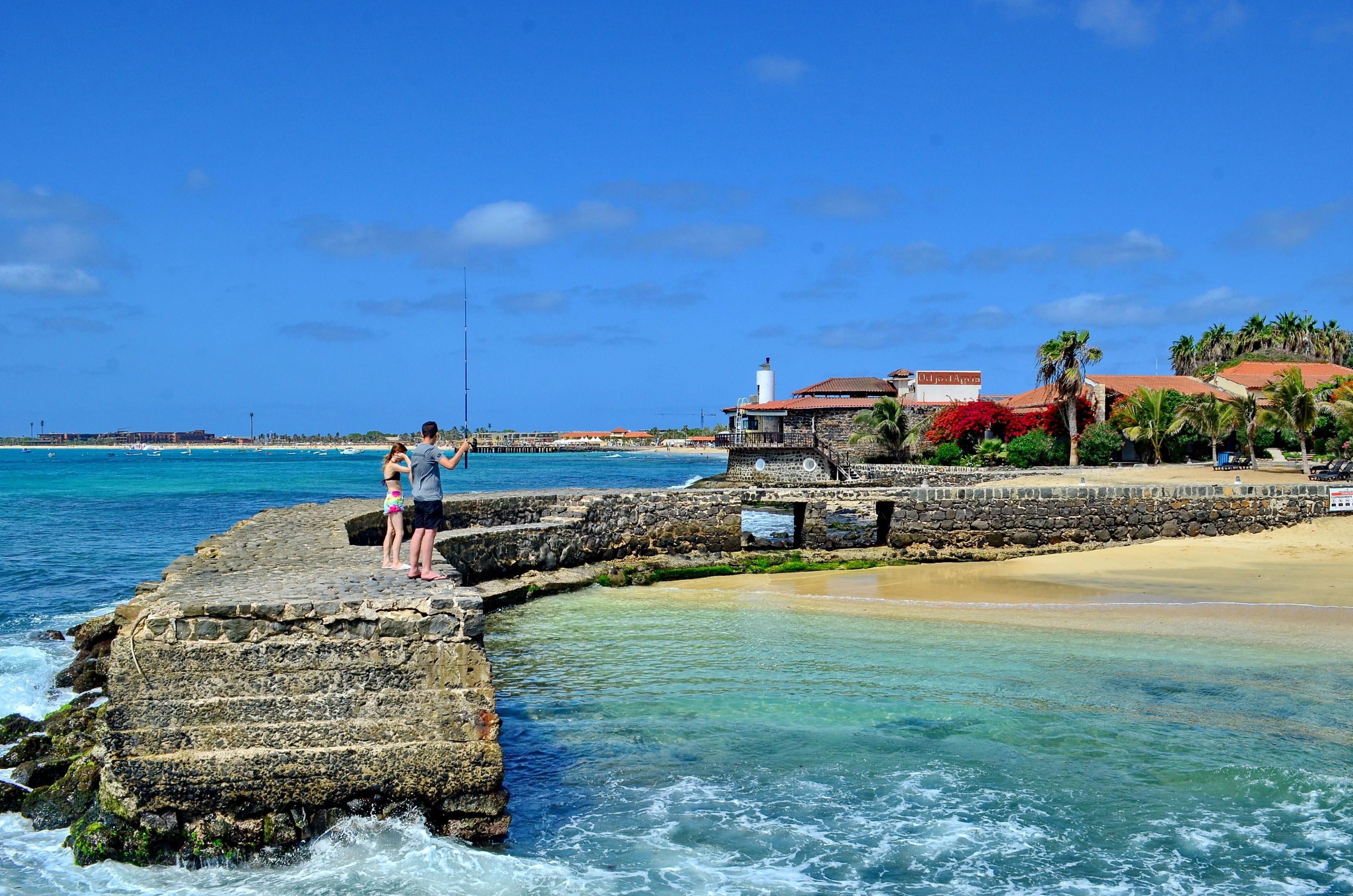 A coastal scene with a stone pier extending into the ocean, a sandy beach, and a building with a red roof in the background.