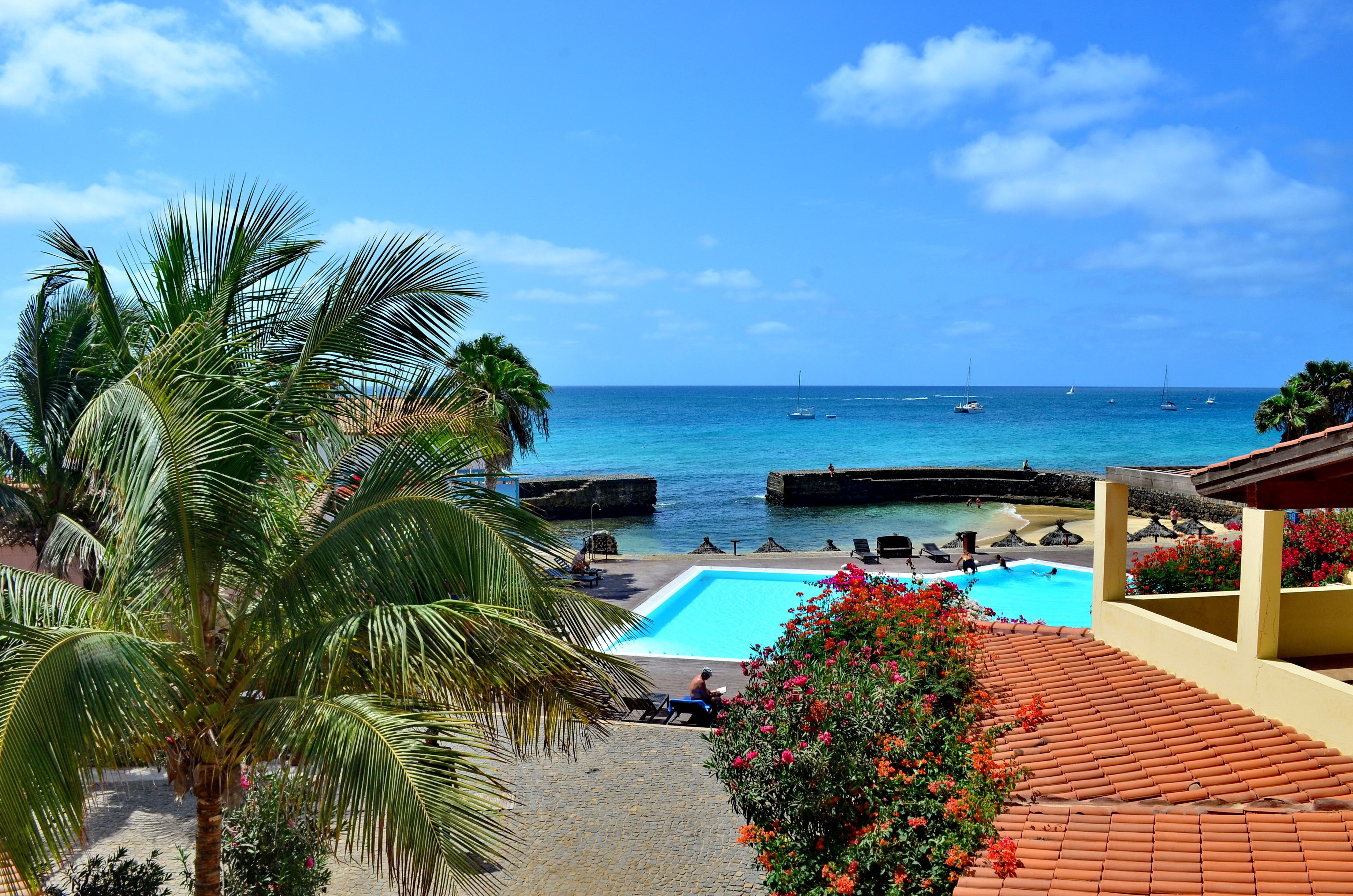 Ein Poolbereich mit Blick auf das Meer und Boote, umgeben von Palmen und einem Ziegeldach.