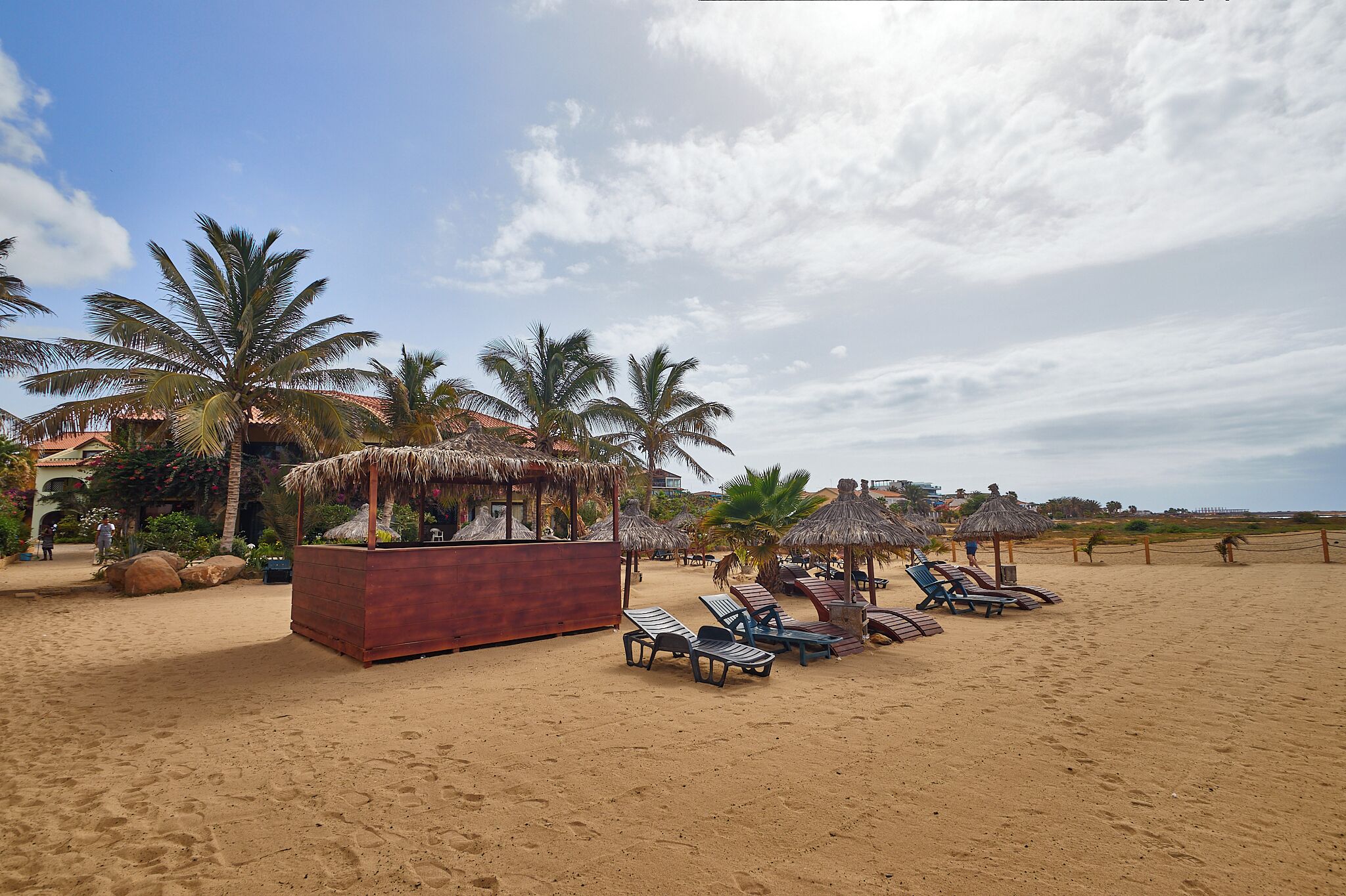 A beach with lounge chairs, palm trees, and a thatched-roof structure.