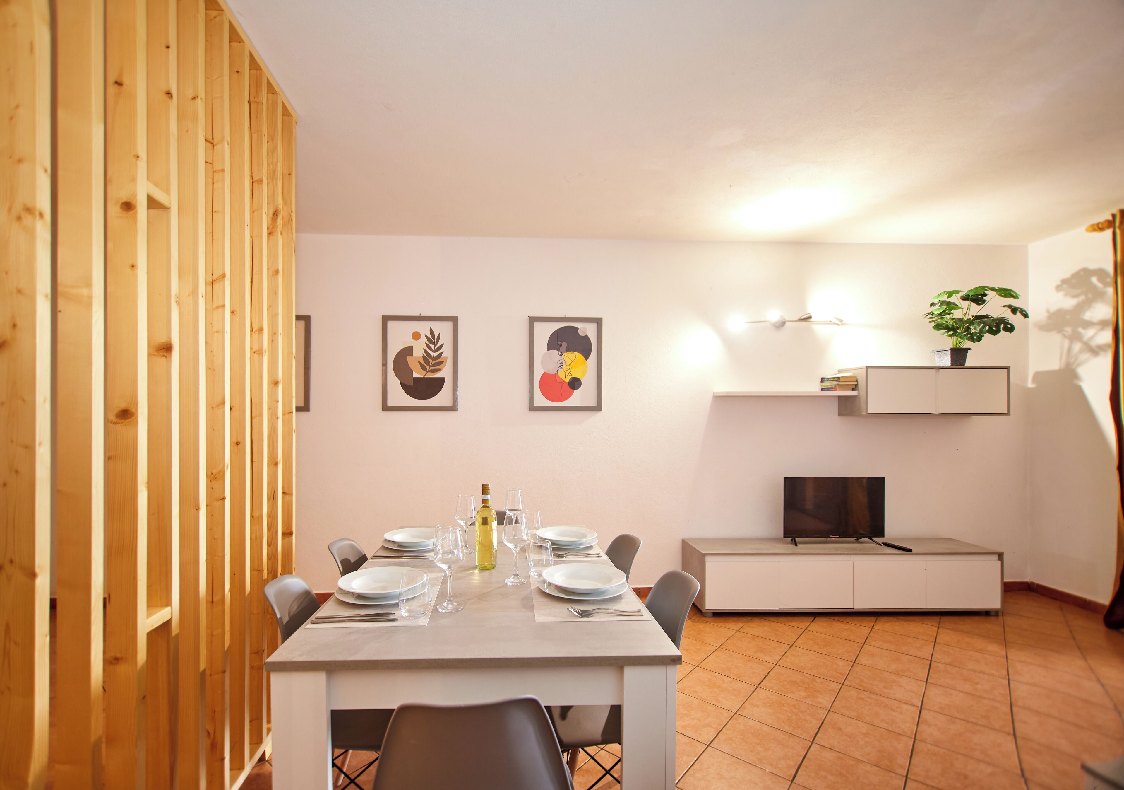 Un comedor con una mesa dispuesta para cuatro, un televisor sobre un mueble bajo y una planta en un estante.