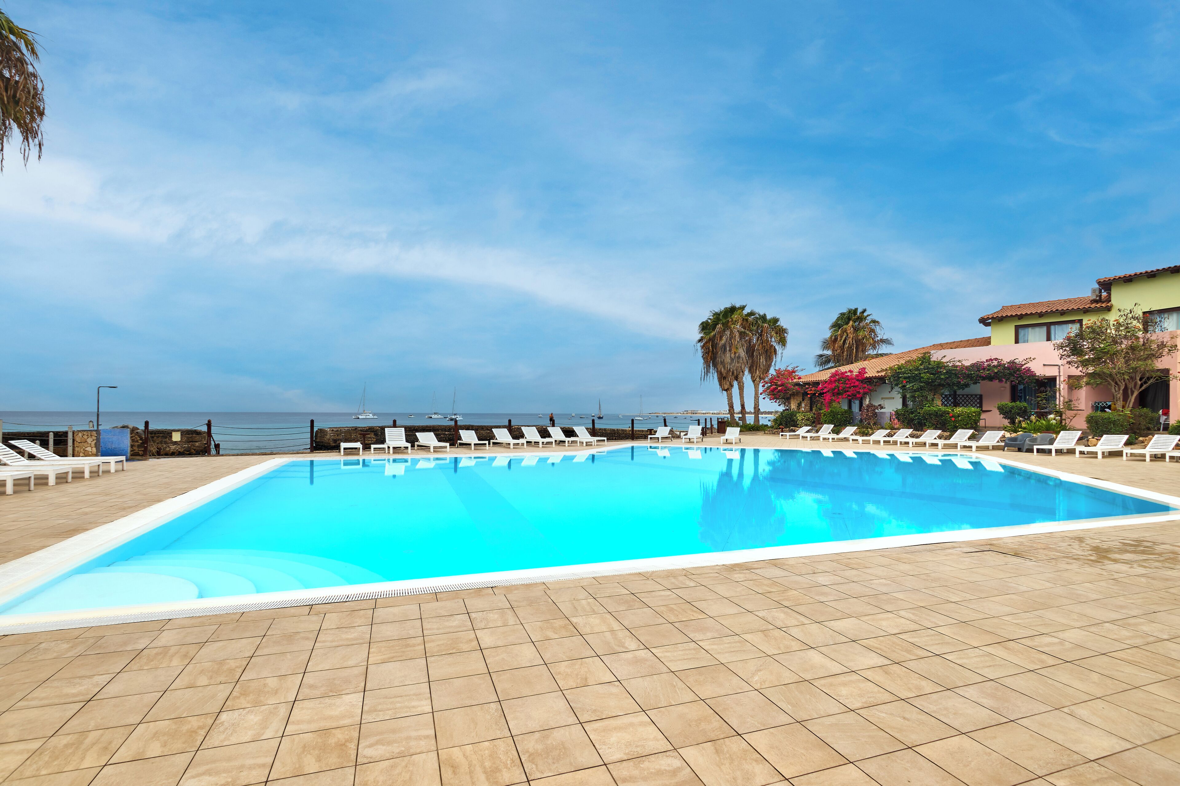 Una piscina rectangular con sillas de descanso blancas a su alrededor, un suelo de baldosas y vista al océano.