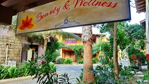 A sign for "Beauty & Wellness" with a flower logo, located in a courtyard with tropical plants and a stone building in the background.