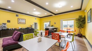 A brightly lit indoor space with yellow walls, multiple tables and chairs, a sofa, a kitchen area, and a potted plant. - Residency Suites - Cotulla (Cotulla)