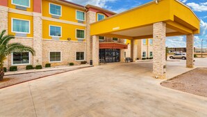 A multi-story hotel with a yellow facade and stone accents, featuring a covered entrance area with a bench and a palm tree. - Residency Suites - Cotulla (Cotulla)