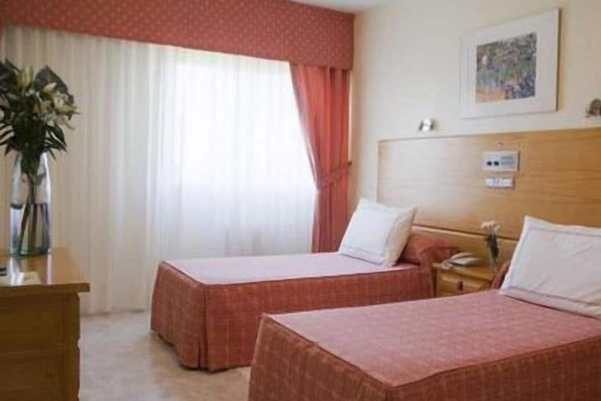 Une chambre d’hôtel avec deux lits, une commode en bois, un vase avec des fleurs, un tableau au mur et une fenêtre avec des rideaux.