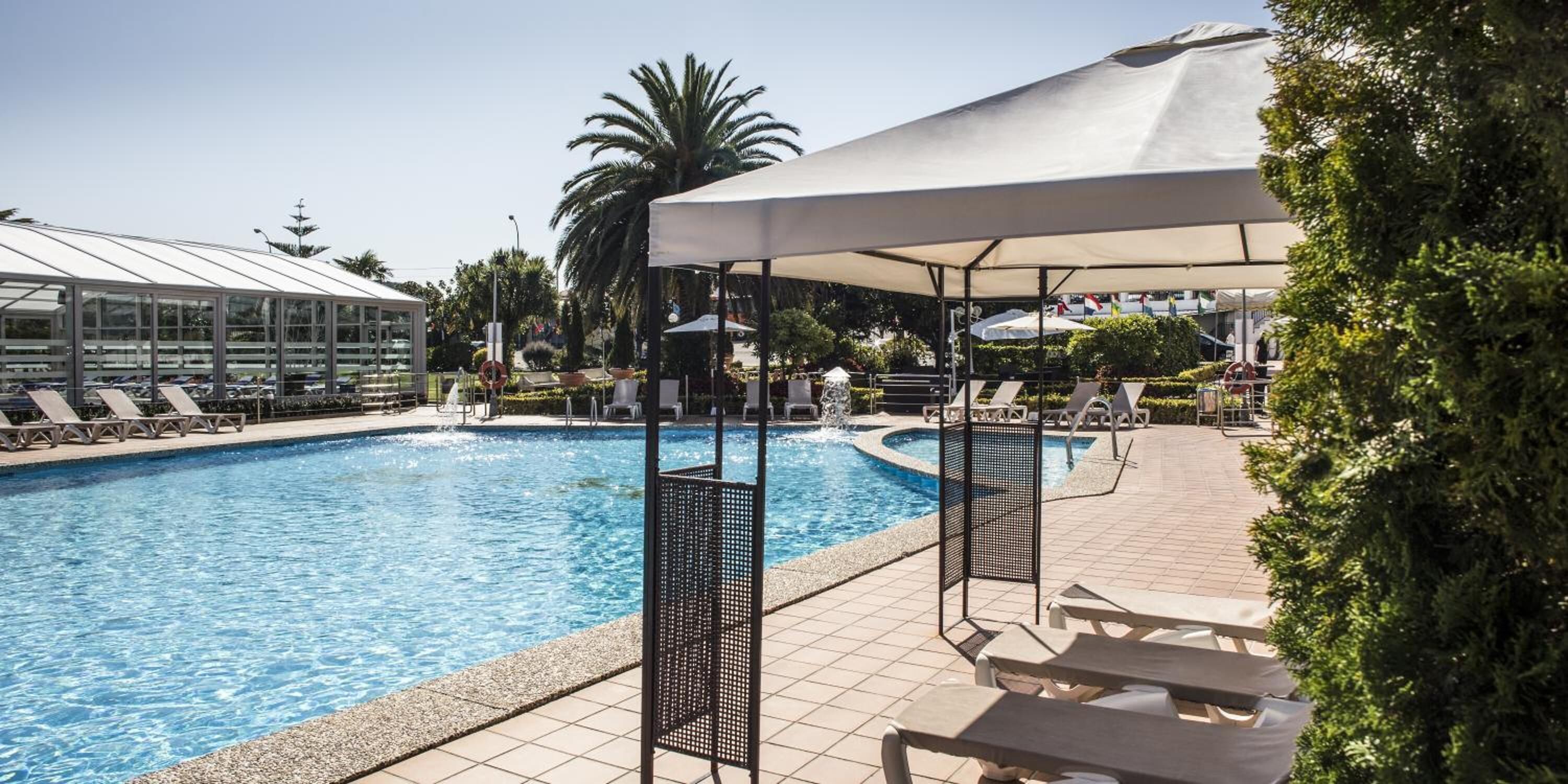 A swimming pool with lounge chairs and umbrellas, surrounded by greenery and a covered area.