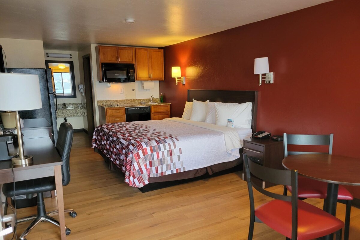 A hotel room with a bed, a desk with a chair, a dining table with a chair, a microwave, and a small kitchenette.