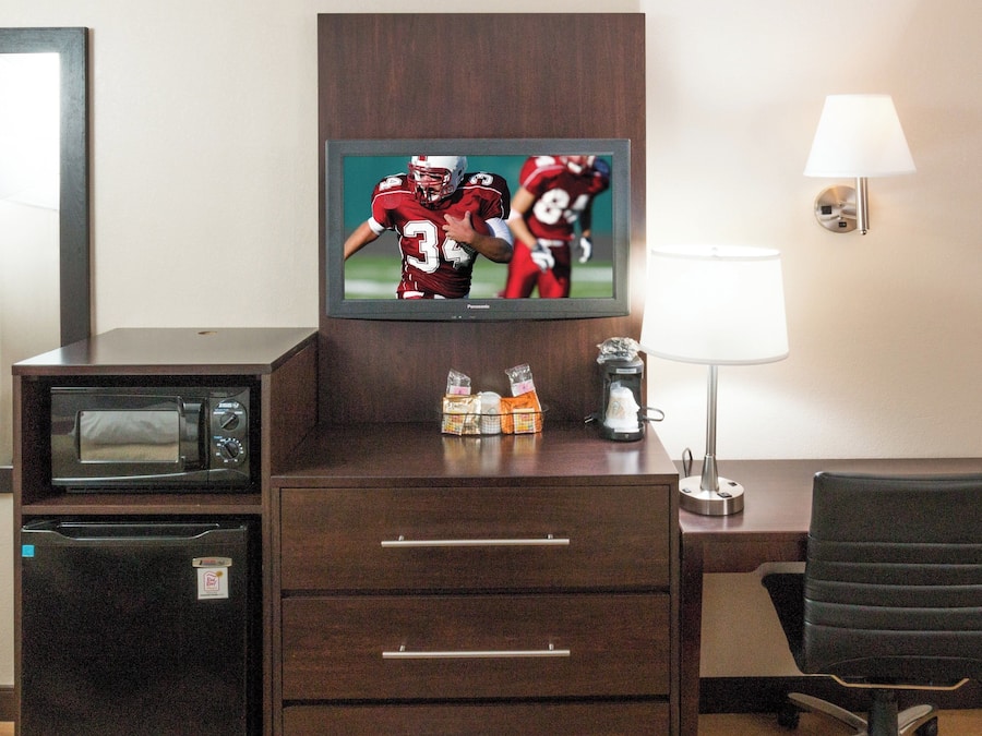 A hotel room with a television displaying a sports game, a desk with a lamp, a chair, and a minibar.