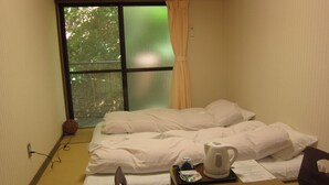Habitación de hotel con una cama, una mesa con hervidor, una silla y una ventana con cortina.